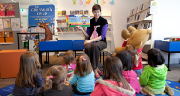 Group of toddlers sitting on a colour rug looking at a teach holding up a picture book and reading out loud