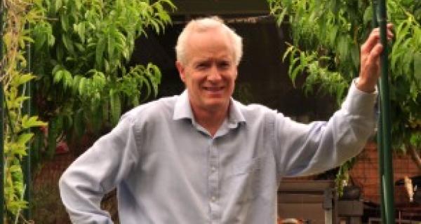 Kenton leans on heavily foliaged garden arch with his Earthship house in the background, proudly displaying his solar panels