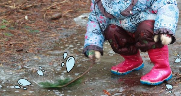 child wearing a blue raincoat and red gumboot playing with a leaf in a muddy puddle