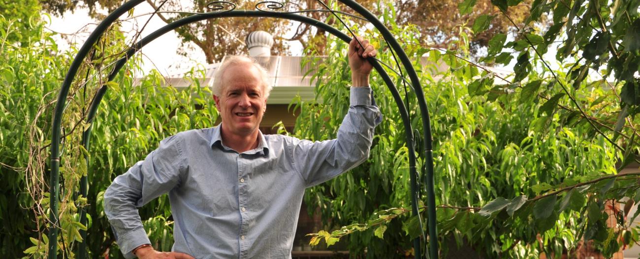 Kenton leans on his garden arch with fruit trees and solar panels on the roof of his Earthship home in the background